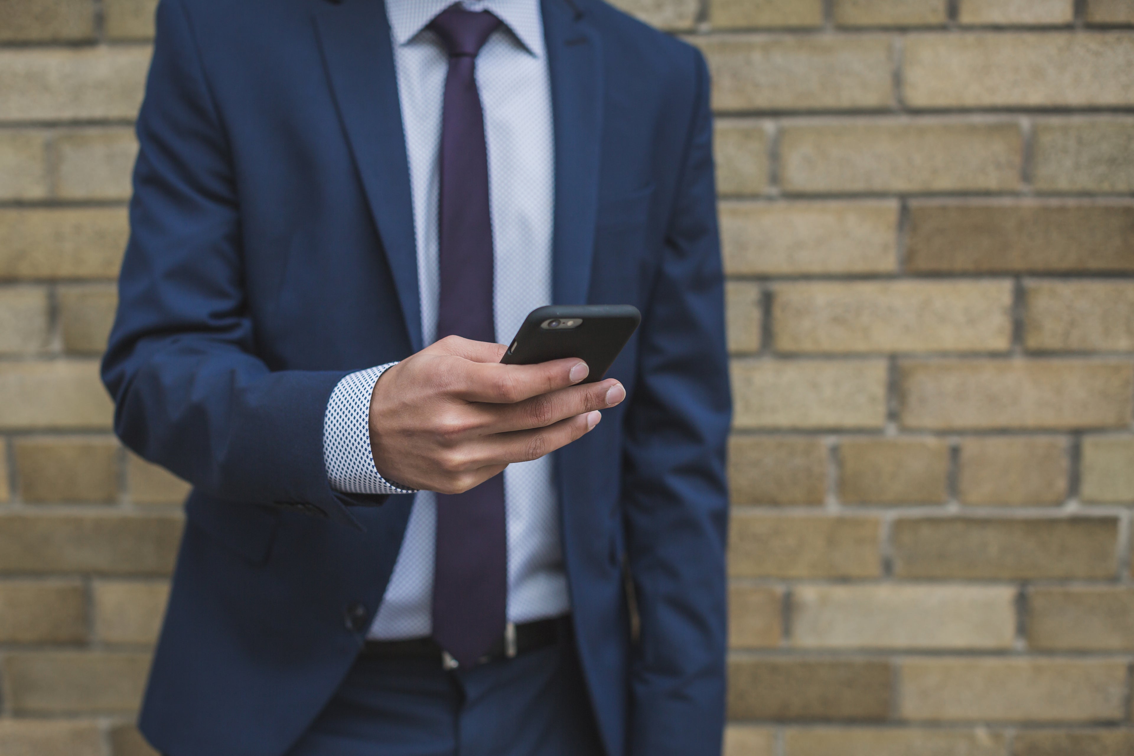 A man in a blue suit and tie stands against a brick wall, holding and looking at a smartphone in his right hand. His head is not visible in the image.