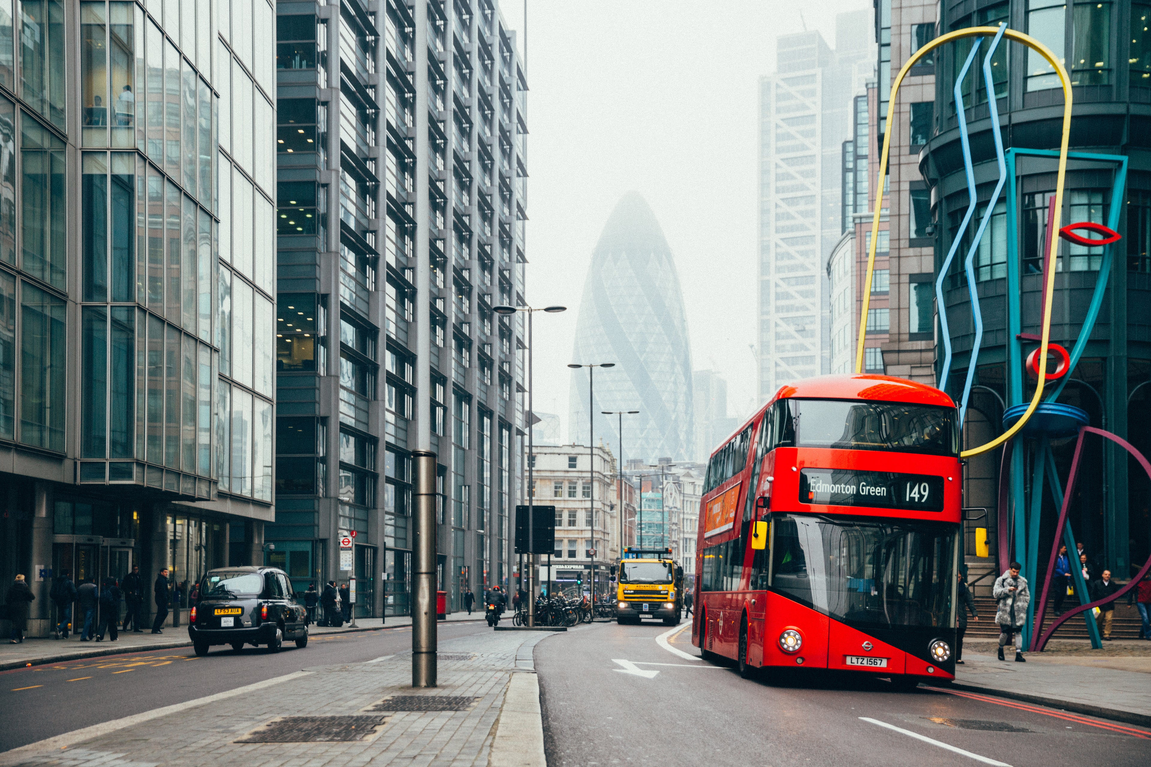 A red double-decker bus labeled “149” drives through a busy London street lined with modern buildings, with the Gherkin skyscraper visible in the background on a cloudy day.